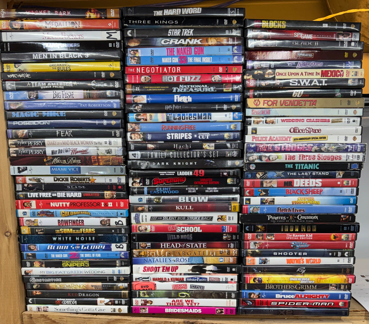 A large collection of DVDs arranged in rows on a wooden shelf, with many spine titles visible. A plywood panel stands to the right, and the shelving is shown in a real indoor setting rather than as a clean studio setup.