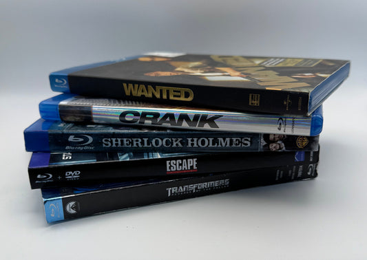 A stack of five physical media cases, including Blu-ray and DVD discs, standing upright on a white surface next to a wooden table. Visible titles include Wanted, Crank, Sherlock Holmes, and Escape.