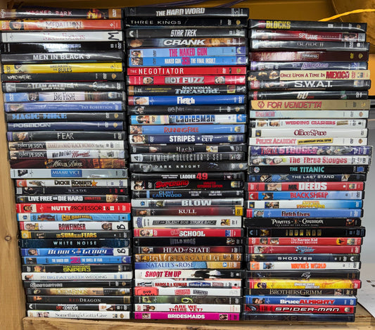 A large collection of DVDs arranged in rows on a wooden shelf, with many spine titles visible. A plywood panel stands to the right, and the shelving is shown in a real indoor setting rather than as a clean studio setup.